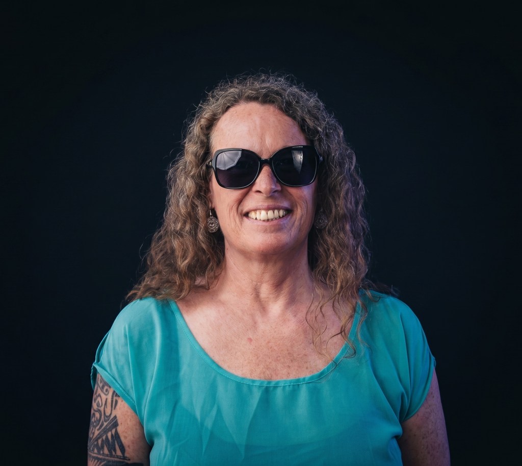 Eine lächelnde Frau mit lockigem, braunem Haar, die eine schwarze Sonnenbrille und ein türkisfarbenes Oberteil trägt, vor schwarzem Hintergrund.