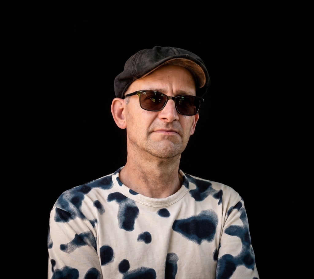 Ein Mann mit Sonnenbrille und einer Mütze steht vor einem schwarzen Hintergrund. Er trägt ein langärmliches T-Shirt mit einem einzigartigen Muster.