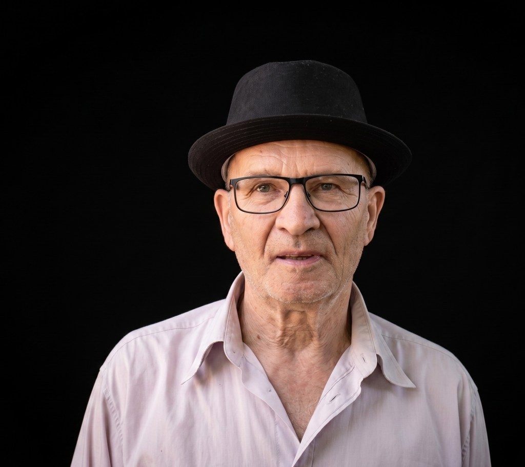 Ein älterer Mann mit einer Brille und einem schwarzen Hut, der in die Kamera blickt. Er trägt ein helles Hemd und hat einen neutralen Gesichtsausdruck. Der Hintergrund ist schwarz.