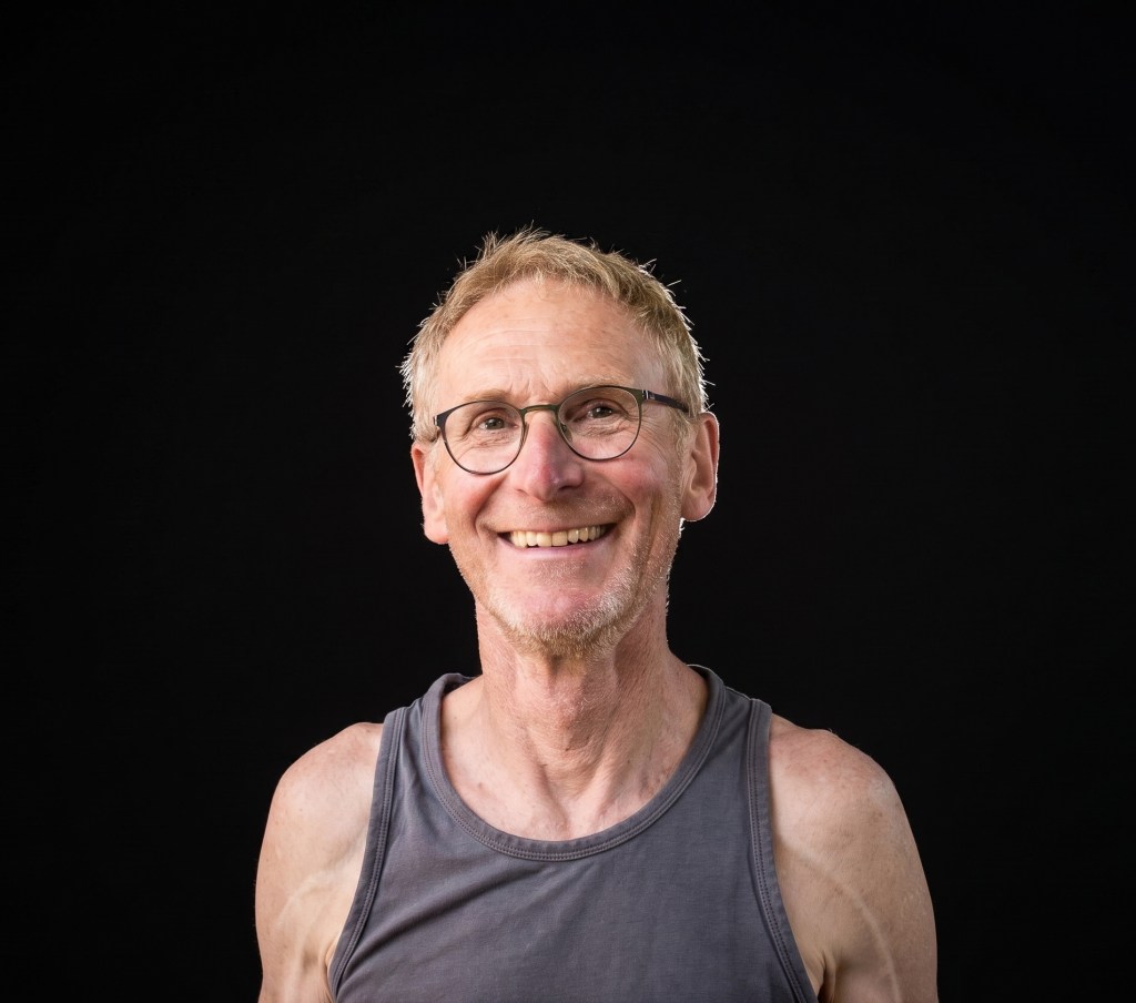 Ein älterer Mann mit Brille und kurzem Haar lächelt freundlich in die Kamera, während er ein graues Tanktop trägt. Der Hintergrund ist schwarz.
