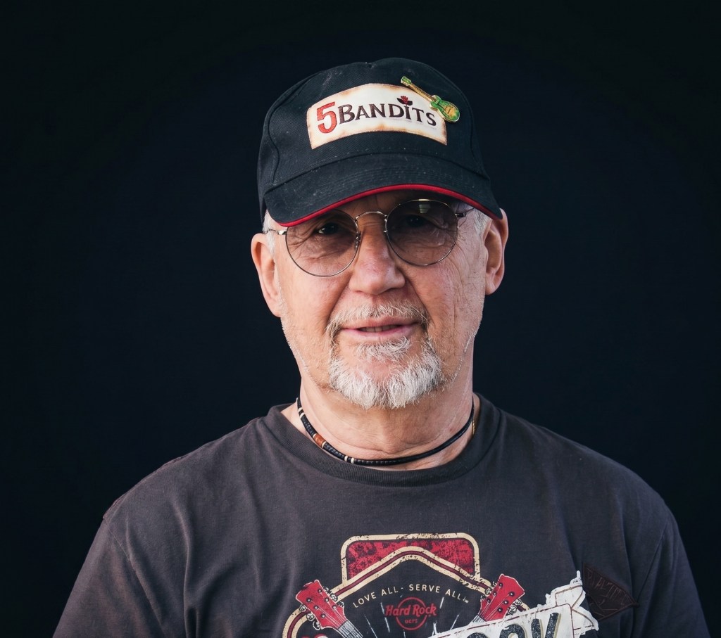 Ein älterer Mann mit einer Halbglatze trägt eine schwarze Kappe mit der Beschriftung '5 Bandits'. Er hat eine Brille auf und einen grauen Bart. Sein T-Shirt hat ein rockiges Design.